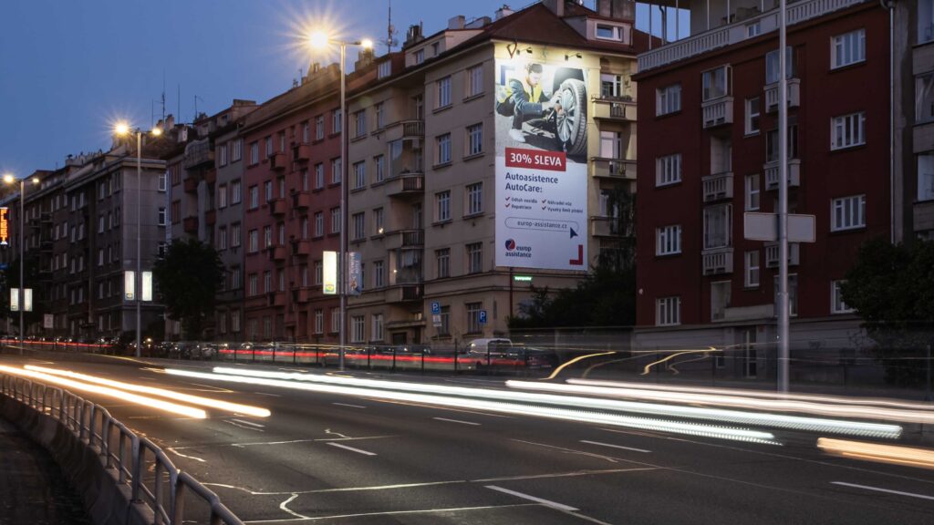 Noční městská scéna zachycující velký reklamní billboard Europ Assistance na boční stěně bytového domu. Billboard zobrazuje reklamu na autoasistenci s 30% slevou a ilustrací mechanika opravujícího kolo. V popředí je zachycena rušná silnice s rozmazanými světelnými stopami projíždějících aut, které vytváří dynamické světelné pruhy. Záběr je pořízen za soumraku, kdy jsou již rozsvícené pouliční lampy, které osvětlují řadu městských bytových domů v různých odstínech červené a béžové barvy.