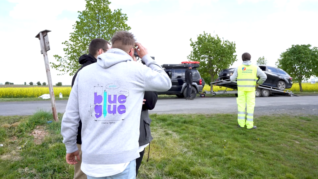 Záběr z natáčení reklamního spotu pro Europ Assistance zachycený v exteriéru. V popředí stojí dva kameramani a zástupce agentury Blueglue v šedé mikině s logem agentury. V pozadí je vidět odtahový vůz s naloženým černým osobním automobilem a pracovník Europ Assistance ve žluté reflexní vestě. Scéna se odehrává na venkovním prostranství s řepkovým polem a stromy v pozadí za zamračeného počasí.