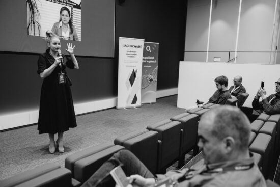 Petra Stupková z Legitas přednáší na konferenci Eshopista, gestikuluje při prezentaci publiku s bannery ACOMWARE a O2.